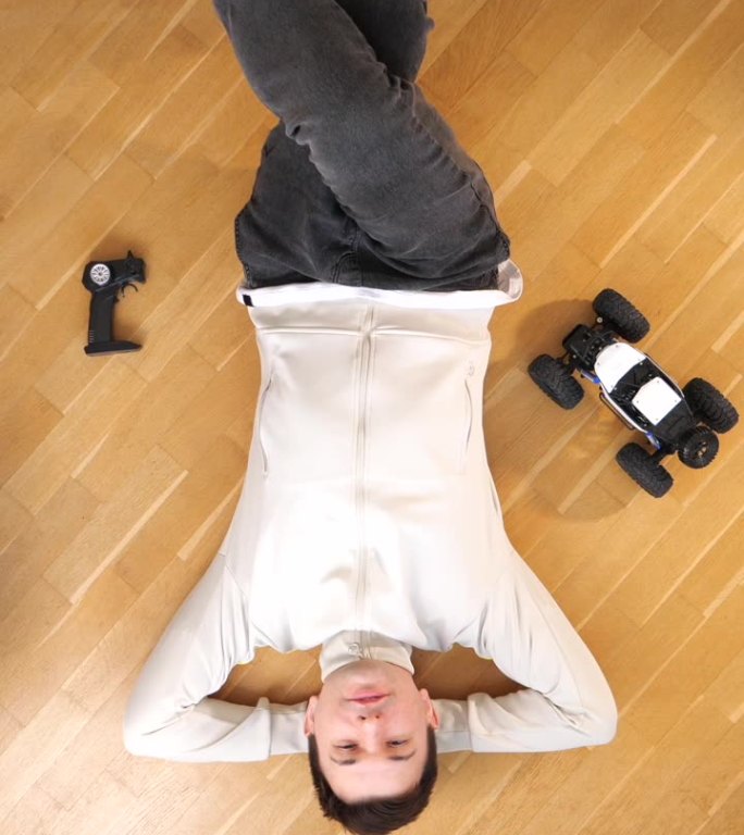 垂直视频男子仰面躺在地板上，遥控汽车模型站在附近