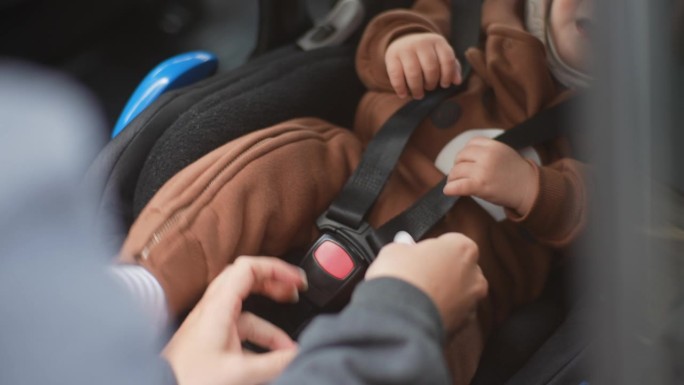 特写镜头中，一位无法辨认的年轻母亲正将安全带系在汽车座椅上的男婴身上。可爱的小孩在车上系上安全带。