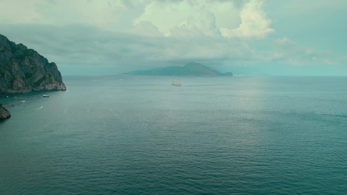 多云的天空下，卡普里岛附近的朦胧海景和帆船。这幅海上场景拍摄了一艘高大的船只在柔和的光线下滑过岛上突