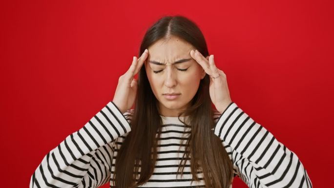 饱受偏头痛折磨的年轻西班牙女性，穿着条纹t恤的美女站在孤立的红色背景下。