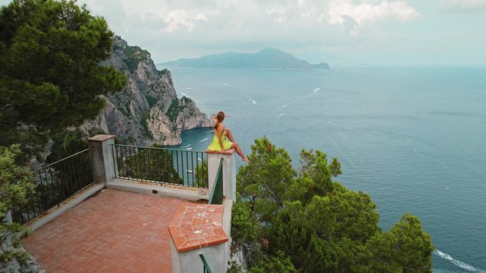 一位身穿黄色连衣裙的妇女坐在风景优美的眺望台上。女孩凝视着宁静的海景，水面上点缀着多艘船。卡普里意大
