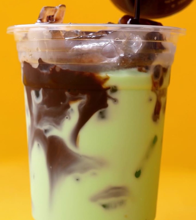 用手将热巧克力倒入装有薄荷绿茶的冰块中，杯底为黄色。
