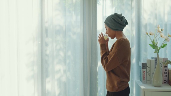 一位年轻的亚洲癌症患者戴着头巾，在家里享受着早晨温暖的阳光和窗边的热饮。与癌症共存。