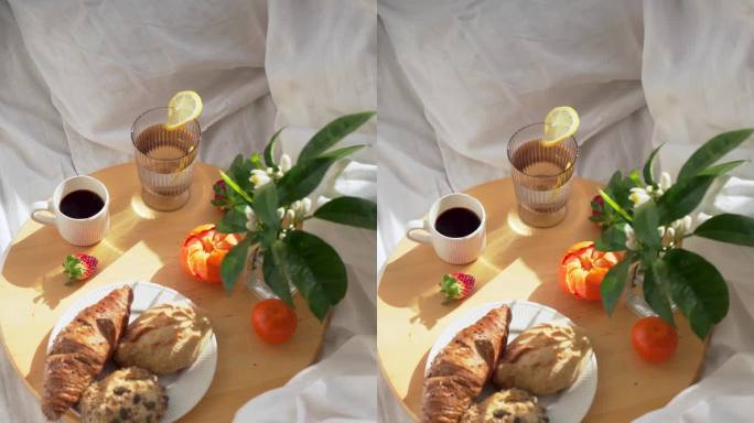 一杯咖啡，橘子和羊角面包放在床上白色亚麻布的圆形木托盘上。垂直4K画面