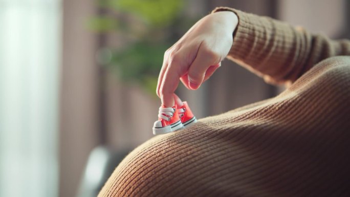 怀孕的孕妇在家里的客厅里穿着鞋在她的大肚子上走着她的手指，怀孕的孕妇在玩她宝宝的鞋子。