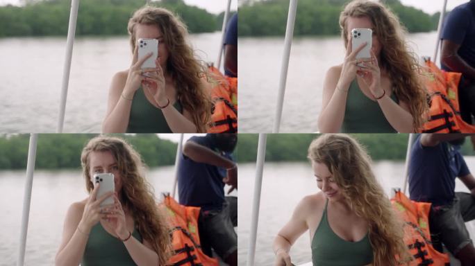 卷发女性在河上巡游时玩智能手机，享受平静的水面。微笑着，在自然环境中使用数字技术。休闲水路之旅放松、