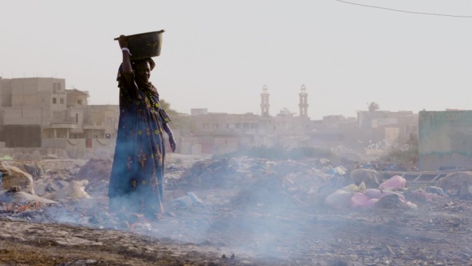 特写镜头。上了年纪的黑人妇女，头上顶着一个塑料桶，里面装着在木屑上熏过的鱼。空气污染。塞内加尔达喀尔