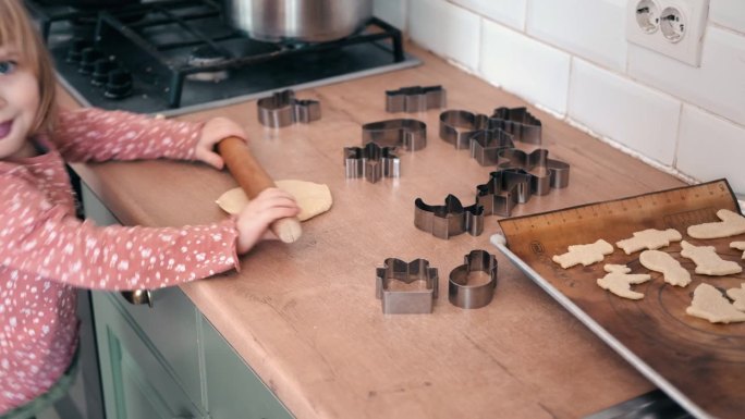 3岁的小女孩站在凳子上，用一根木制擀面杖在厨房的台面上擀出饼干面团。金属烘烤模具。童年烘焙的乐趣，家