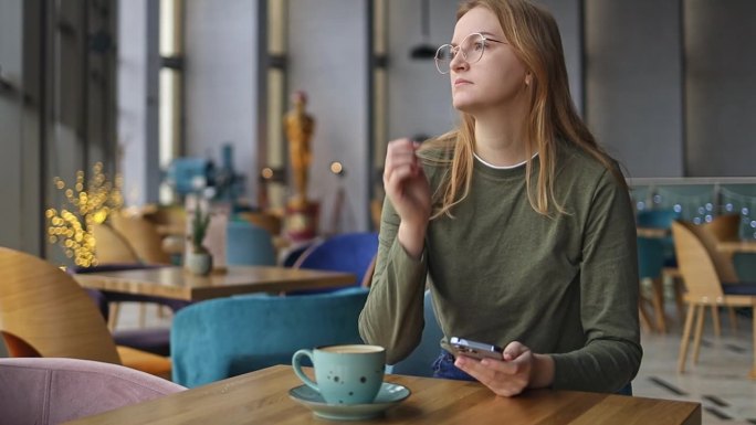 年轻漂亮的30多岁的金发女子，戴着眼镜，坐在咖啡馆里拿着手机看智能手机。快乐的女孩用手机和眼神调情。