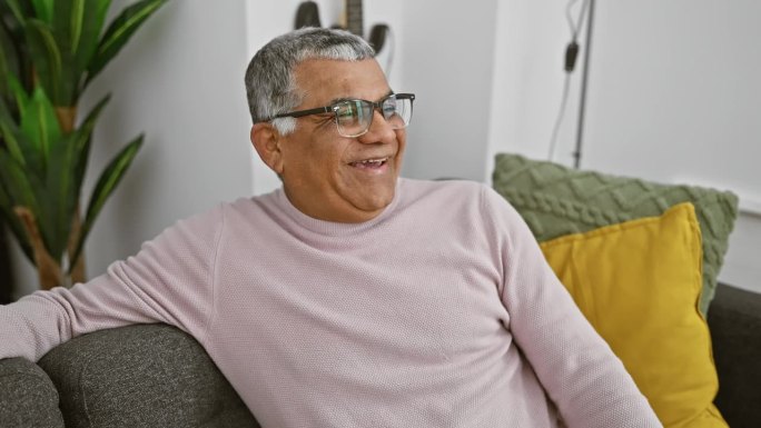 在舒适的客厅里，一个白发苍苍的男人坐在灰色的沙发上微笑着，沙发上有鲜艳的靠垫，背景是一棵植物。