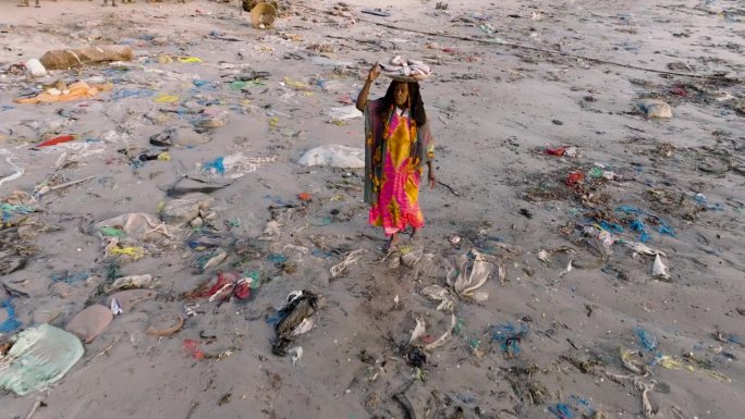 空中跟踪特写。在塞内加尔的达喀尔，一名卖鱼的黑人妇女头上顶着一个篮子，穿过鱼市海边可怕的塑料污染
