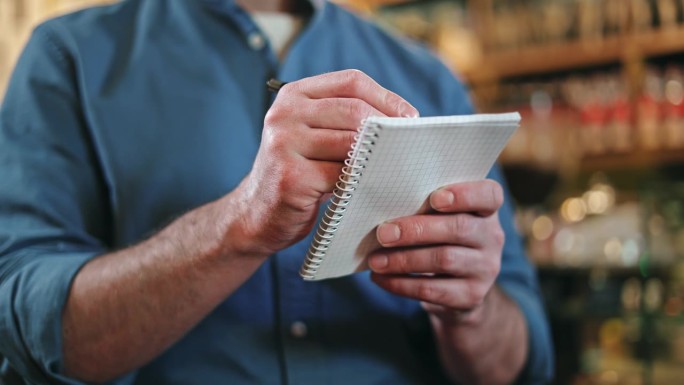 一名穿着蓝色衬衫、卷起袖子的男性餐饮服务人员在室内在纸质笔记本上写订单。有能力的服务员在餐厅做记录。