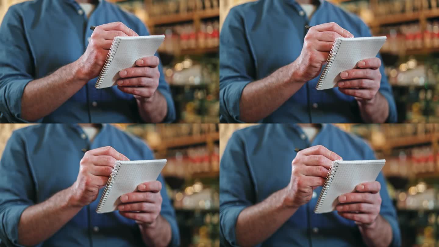 一名穿着蓝色衬衫、卷起袖子的男性餐饮服务人员在室内在纸质笔记本上写订单。有能力的服务员在餐厅做记录。