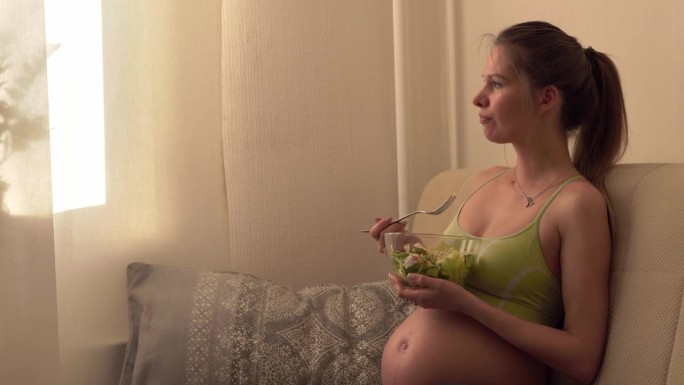 为了不发胖，躺在沙发上露着肚子吃沙拉的孕妇
