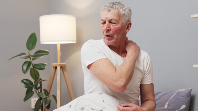 成熟的白人男子在不舒服的睡眠后坐在床上处理身体问题，摸着脖子，表达脖子疼痛带来的不适