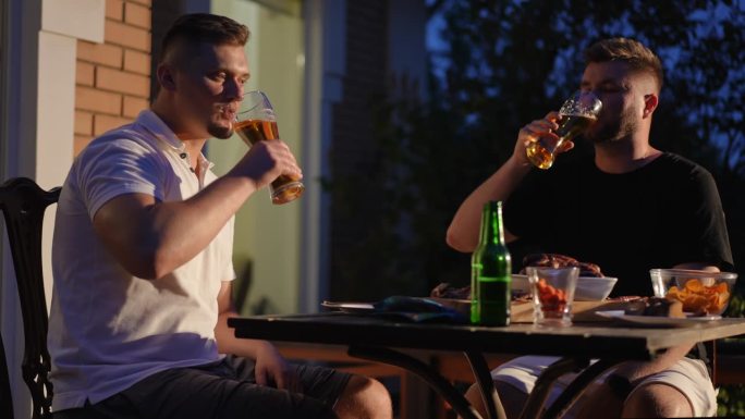 夏日的夜晚，两个男人端着啤酒坐在户外的木桌旁，桌上放着烤香肠和薯条。男人们碰杯喝啤酒。