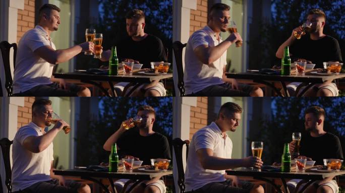 夏日的夜晚，两个男人端着啤酒坐在户外的木桌旁，桌上放着烤香肠和薯条。男人们碰杯喝啤酒。