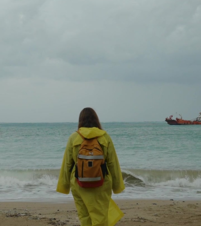 岩石间的海湾，远处的一艘船，一个穿着黄色雨衣的女人迎着风沿着沙滩走。多云的天气。