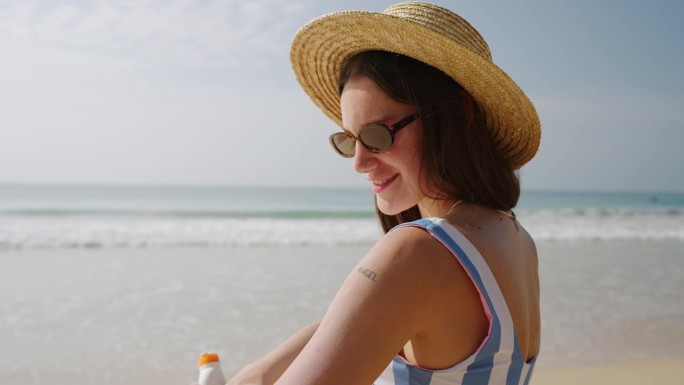 快乐的年轻女子在海边涂抹防晒乳液。戴草帽的漂亮女孩在皮肤上涂防水防晒霜以防止紫外线。穿着比基尼的女士
