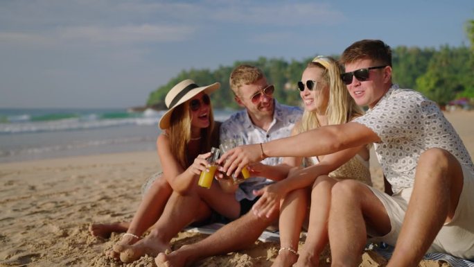 四个快乐的朋友坐在沙滩上，喝着康普茶，在热带之旅中欢呼和聊天。两对浪漫的情侣笑着一起放松，在海边喝着