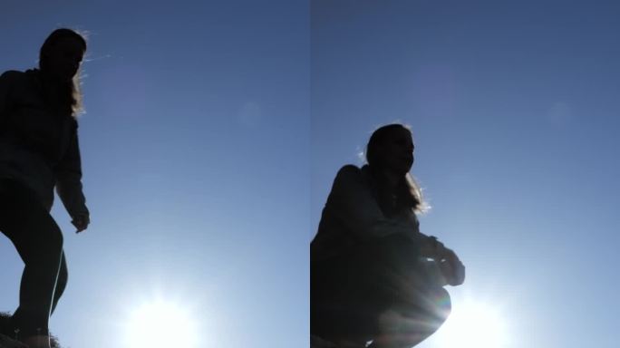 一个女孩的剪影在蓝天和日落的背景下，坐在山顶上，举起双手，在大自然的怀抱中感受自由和快乐。垂直。