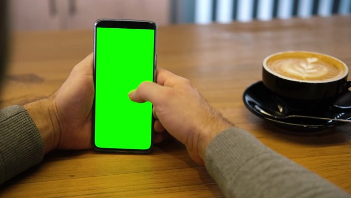 一个年轻人坐在咖啡馆里，手里拿着绿色的智能手机模型屏。男性使用色度键手机。垂直模式。触摸，滑动显示屏
