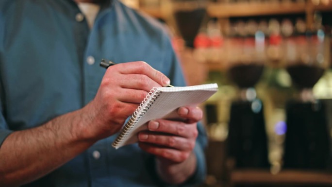 一个穿着蓝色衬衫，卷起袖子的男人在复古风格的咖啡馆工作时用钢笔在抄写本上写字的特写。专业男服务员记录