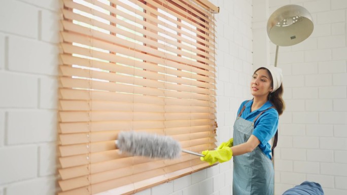 亚洲管家或女佣用鸡毛掸子清洁房子客厅的窗帘或窗帘，她在工作时看起来很开心。