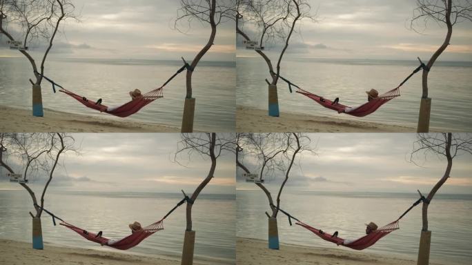 一个穿着浅色衣服的年轻人躺在海边树木之间的吊床上。沙滩，大海，多云的天空为背景。