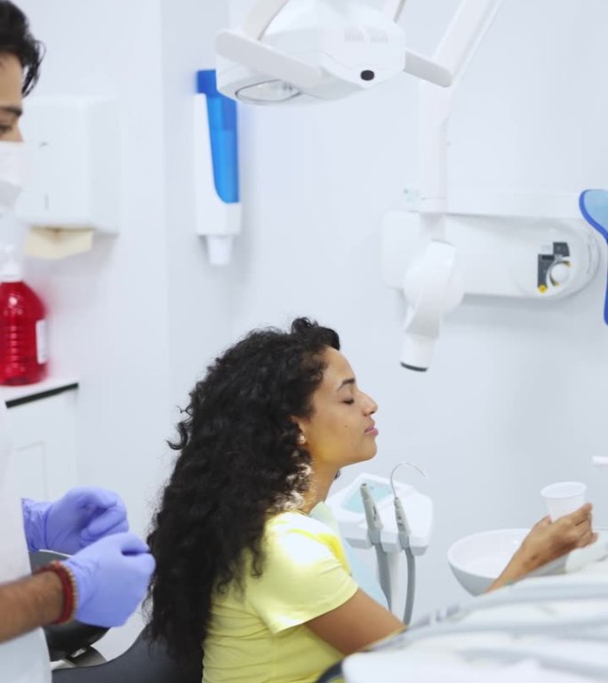 垂直视频的一个年轻的拉丁妇女病人冲洗她的口腔在牙槽，而她的牙医正在等待继续程序。