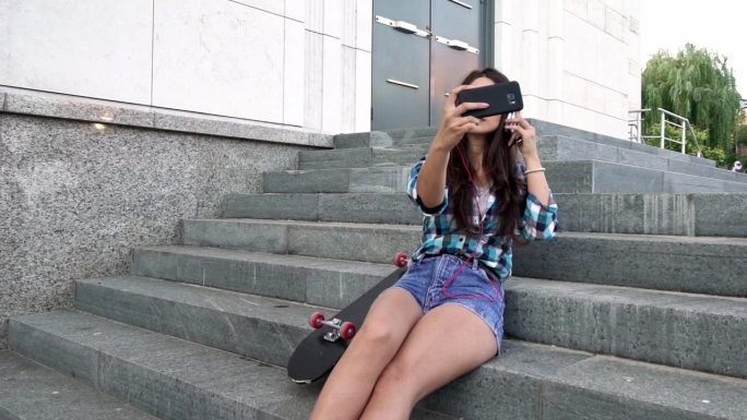 年轻的潮女手拿滑板坐在城市的台阶上自拍