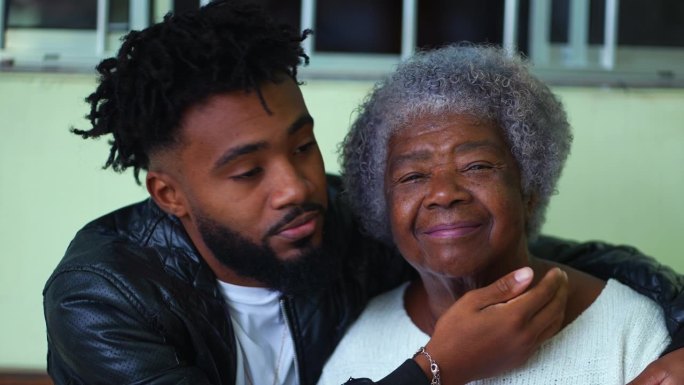 20多岁的年轻非裔美国孙子亲吻年迈的祖母的脸颊，深情地拥抱着家中的长辈，温柔的代际纽带