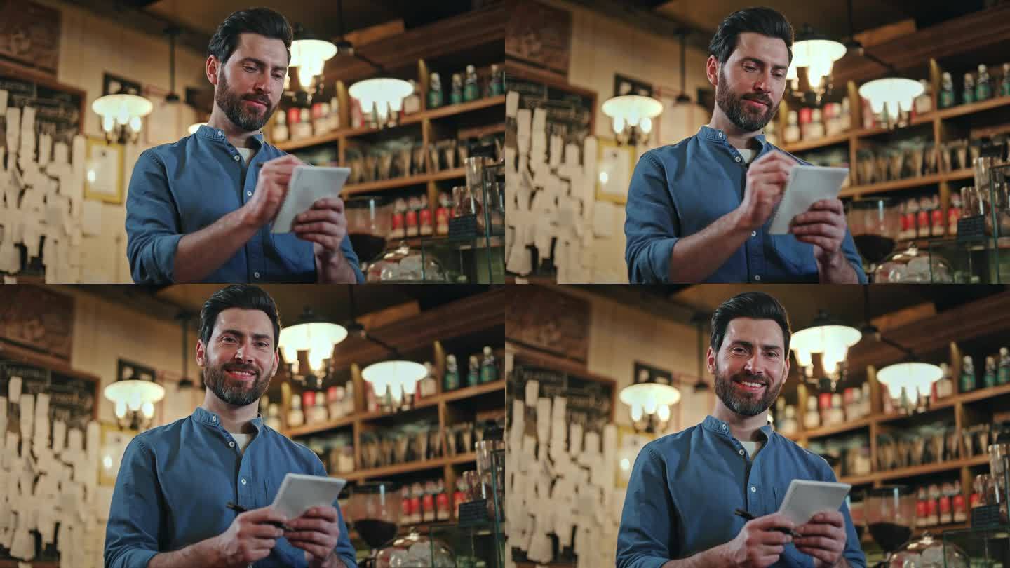 餐馆老板站在摆满酒瓶的架子上写着购物清单。穿着蓝色衬衫的白人大胡子男子，脸上带着积极的表情看着镜头。