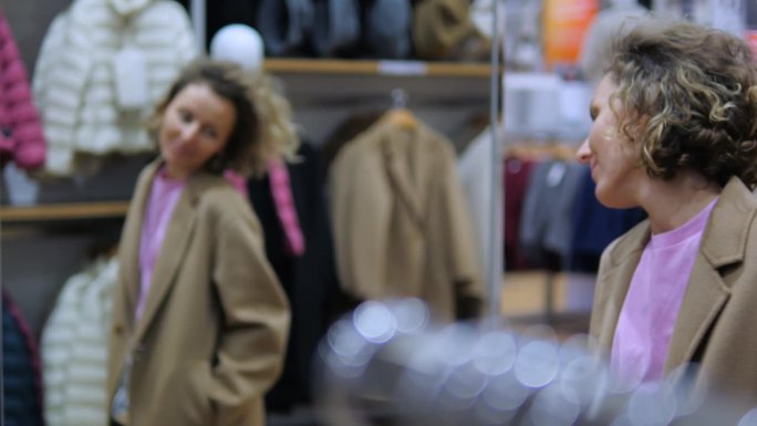 年轻女子在服装店试穿时髦的大衣。时尚的购物者在精品店的镜子前检查她选择的衣服。购物狂喜欢在打折季买衣