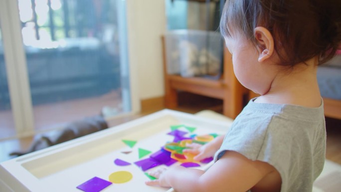 一个蹒跚学步的孩子在光桌上玩半透明的彩色形状，展示了一个明亮的、吸引人的活动，刺激感官和认知的发展