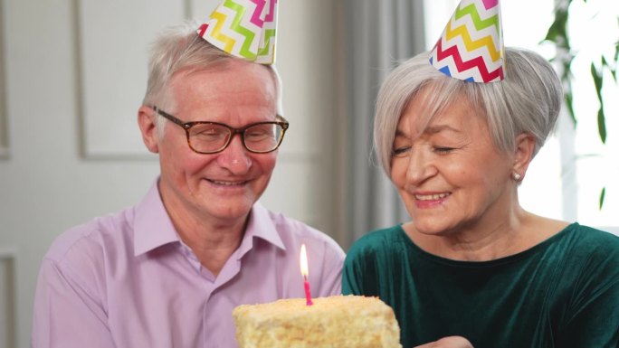 许个愿。家庭高级夫妇戴着派对帽在家里一起庆祝生日纪念日。老人吹灭了生日蛋糕上燃烧的蜡烛。老太太祝丈夫