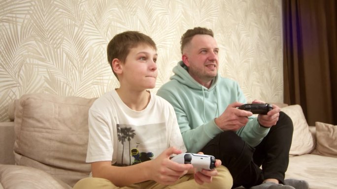 一位父亲和他的小儿子坐在沙发上玩游戏机。周末活动，空闲时间，家庭娱乐和视频游戏的概念。