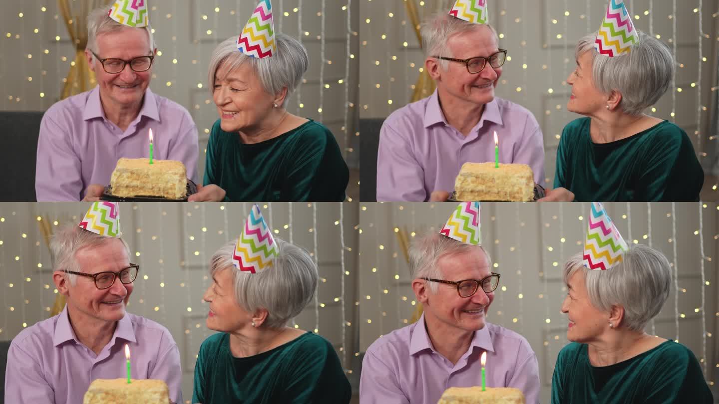 许个愿。家庭高级夫妇戴着派对帽在家里一起庆祝生日纪念日。老太太吹灭了生日蛋糕上燃烧的蜡烛。老人祝妻子