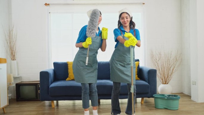 两个亚洲女管家或女佣站在一起，拿着清洁房子的工具，快乐地跳舞。