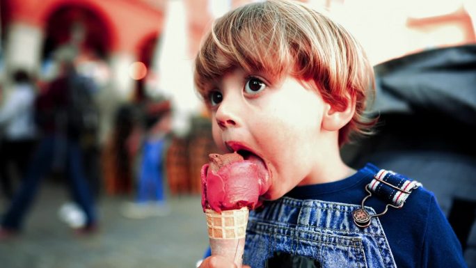 一个小男孩坐在婴儿车上吃冰淇淋甜筒，背景是模糊的行人。凌乱的金发小孩吃清爽点心的特写