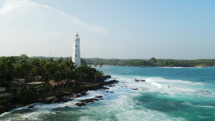 无人机拍摄的热带海岸旧灯塔，巨浪冲击着棕榈树附近的岩石海岸。白塔巍然屹立，碧海环抱海湾名胜古迹。