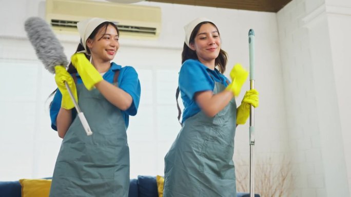 下图两个亚洲女管家或女佣站在一起，拿着清洁房子的工具，快乐地跳舞。