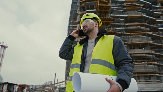 身穿防护服的年轻男建筑师在未完工的建筑前拿着蓝图接电话