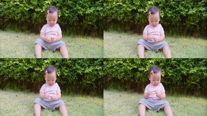 一个蹒跚学步的小女孩坐在草地上，手里拿着一片叶子。画面平静祥和，宝宝在户外玩耍