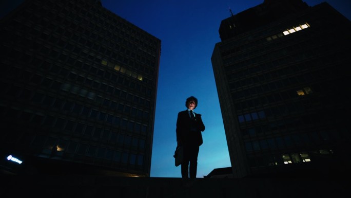 在黑暗的城市里，一个拿着智能手机的商人在大楼下面。思考下一个大生意。