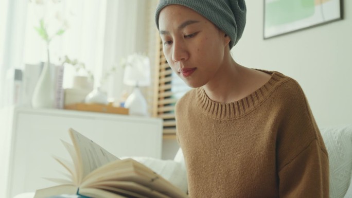 一名年轻的亚洲癌症患者，戴着头巾，在阳光明媚、植物丛生的客厅里，坐在舒适的沙发上，喝着咖啡，读着书。