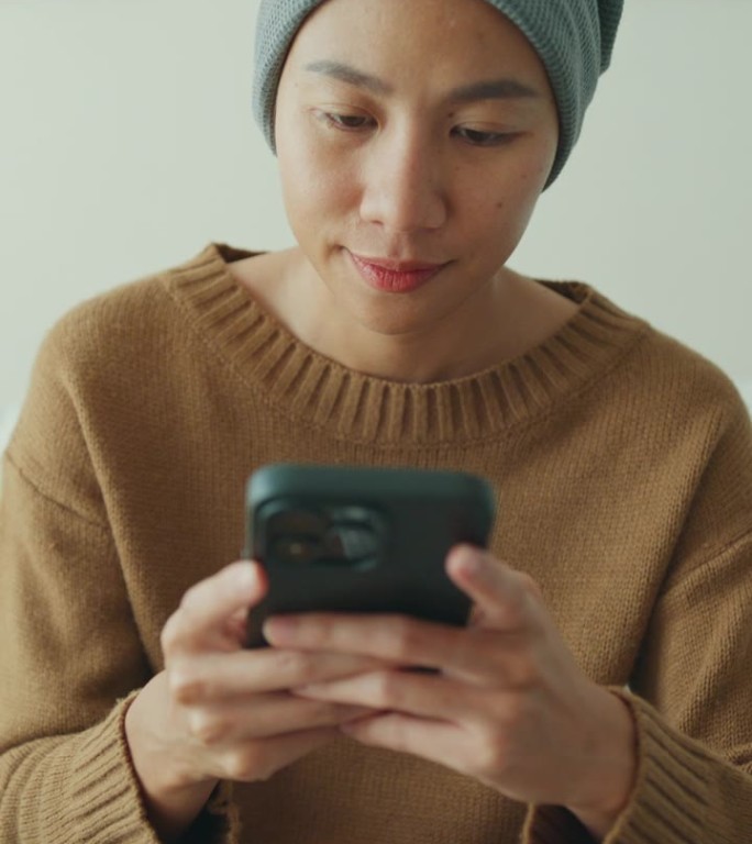 一位年轻的亚洲癌症患者穿着便服，戴着头巾，坐在舒适的沙发上玩手机，周围是一个明亮的，充满植物的客厅。
