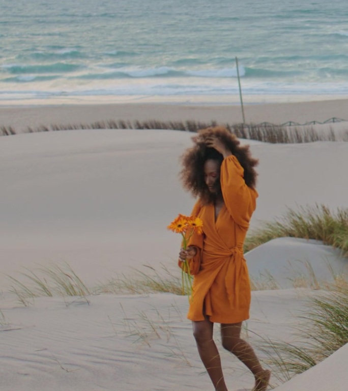 女人走在沙滩黄昏与橙色的花垂直。捧花女孩