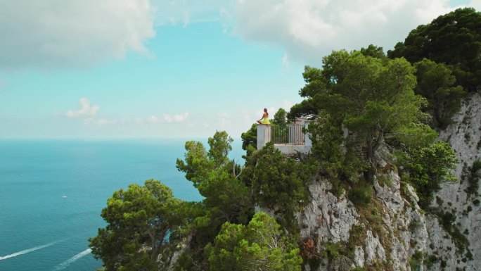 女子在充满活力的悬崖绿洲俯瞰大海，以莲花的姿势冥想。阳光洒在卡普里岛的悬崖上，展现了大自然真正的辉煌