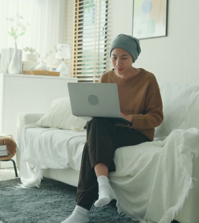 一名年轻的亚洲癌症患者，戴着头巾，坐在一间光线充足、植物装饰的房间里的沙发上，舒舒服服地摆弄着笔记本